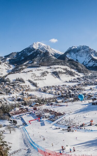 Skirennstrecke und Ortschaft im Vordergrund und blauer Himmel, verschneite Wälder und Berge im Hintergrund. | © Harald Wisthaler