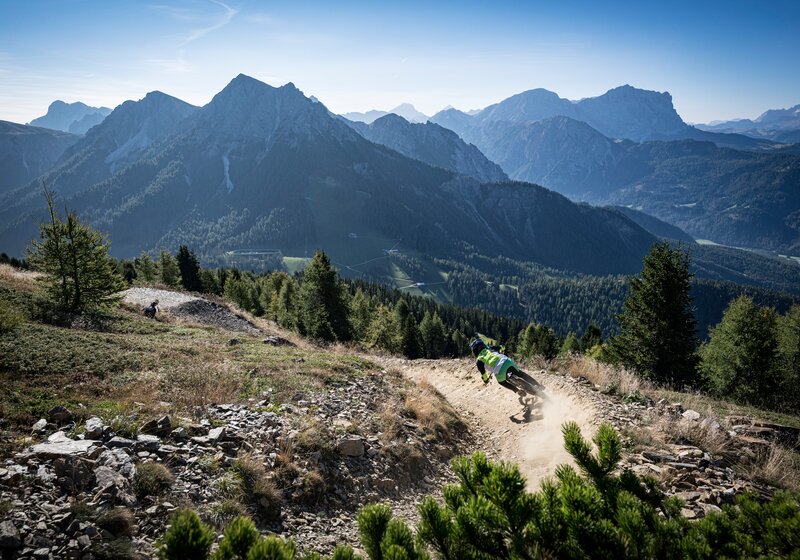 Eine Person fährt eng in die Kurve gelehnt auf einem Mountainbike einen Bergpfad entlang, im Hintergrund das Bergpanorama und strahlend blauer Himmel. | © Stromberg - Enduro Tirol
