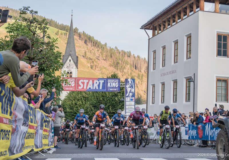 Der Startschuss fällt – zahlreiche Radsportler starten dicht gedrängt in ein Rennen vor einem Gebäude mit Kirchturm. | © Gianvito Coco