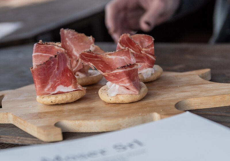 Frisch geschnittener Speck wird auf einem Holzbrett angerichtet, daneben liegt ein Infoflyer. | © Gianvito Coco