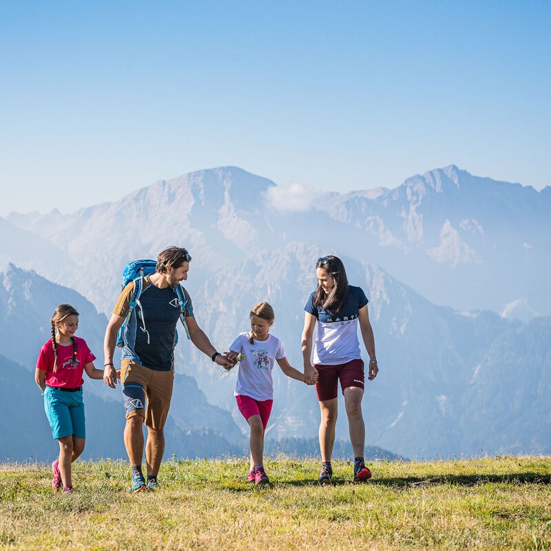 Ein Mann, eine Frau und zwei Kinder laufen auf der Bergwiese am Kronplatz-Gipfel Richtung Kamera mit den Dolomiten im Hintergrund. | © Harald Wisthaler