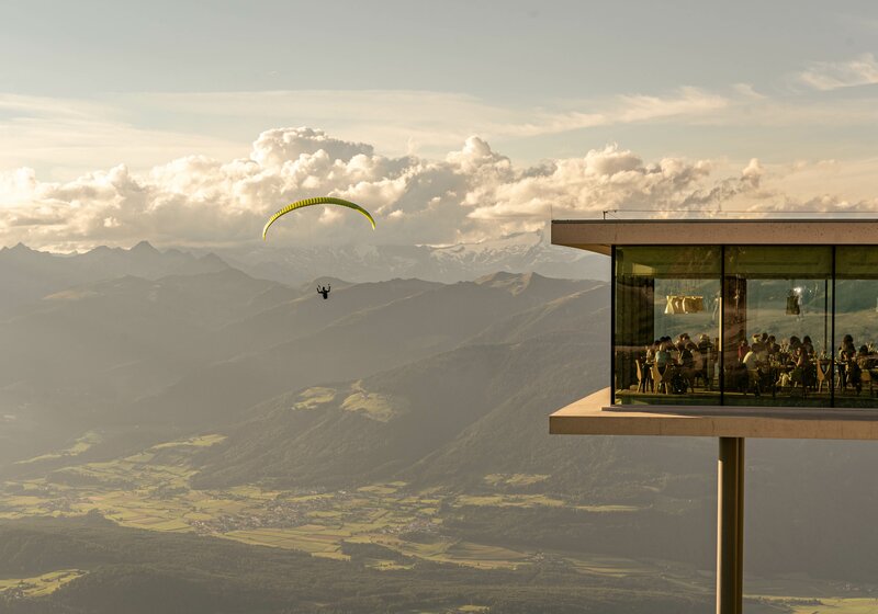 Ein Paragleiter fliegt neben dem AlpiNN Restaurant bei Sonnenuntergang. | © Skirama Kronplatz