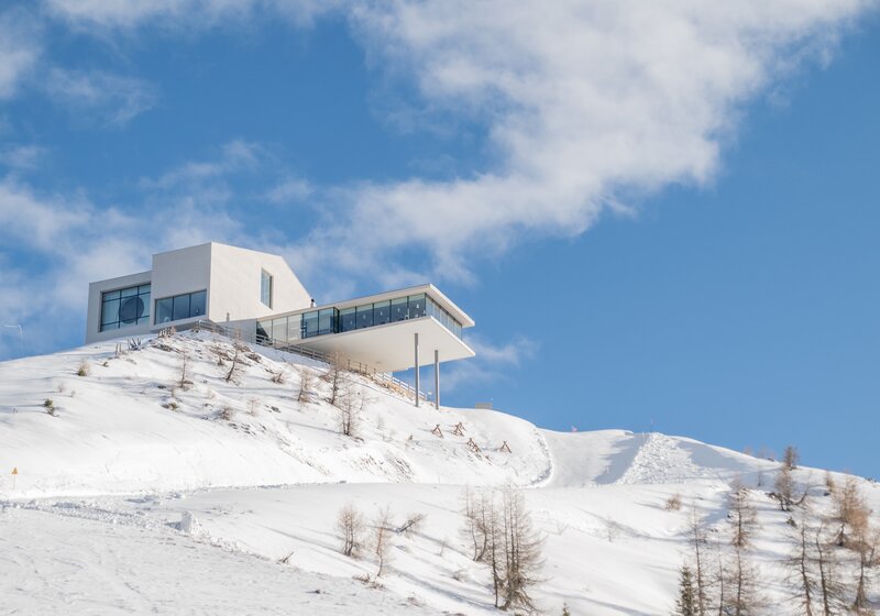 Ein modernes weißes Gebäude thront auf einem verschneiten Hügel mit Skipiste. | © Gianvito Coco