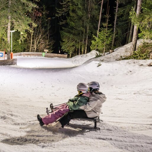Zwei Menschen rodeln lachend eine schneebedeckte, kurvige Waldpiste hinab. | © Gianvito Coco