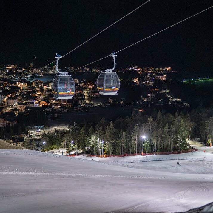 Zwei Gondeln schweben nachts über eine hell ausgeleuchtete Skipiste mit Blick auf eine Stadt im Tal. | © Gianvito Coco